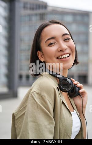 Foto verticale di una bella donna asiatica che si posa con le cuffie intorno al collo, sorridente e ridente, in piedi per strada alla luce del giorno Foto Stock