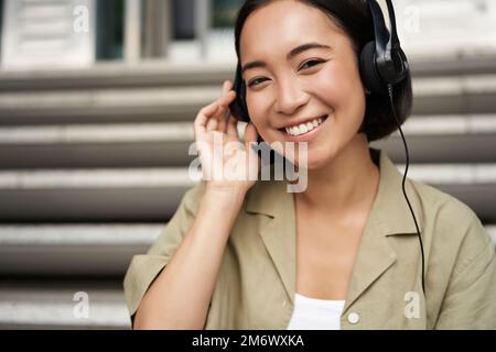 Felice ragazza asiatica ascolta la musica in cuffie, sorride, siede all'aperto sulle scale Foto Stock