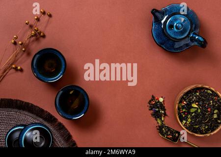 Teiera blu, tazza di tè e foglie di tè su sfondo marrone. Tè alle erbe caldo è nella teiera sul tavolo. Spazio di copia. Foto Stock