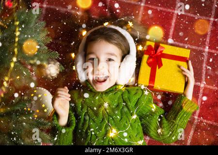 Ragazza in un caldo maglione lavorato a maglia e cuffie in pelliccia è sdraiata su una coperta vicino all'albero di Natale con una scatola regalo. Vigilia di Natale, Foto Stock