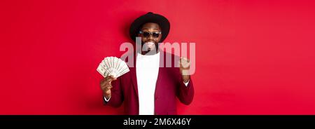 Ragazzo afro-americano allegro in occhiali da sole, cappello nero e blazer, vincendo denaro premio, tenendo dollari e cercando soddisfatto, s Foto Stock