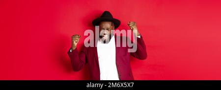 Uomo nero riuscito in vestito che dice sì, gioendo di vincere o di raggiungere l'obiettivo, alzando le mani in su e gridando di gioia, indietro rosso Foto Stock