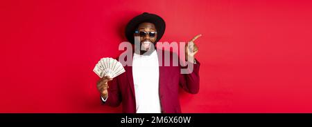 Bell'ed elegante uomo nero che punta le dita a sinistra mentre mostra i soldi, tenendo i dollari e dimostrando il logo, in piedi sopra Foto Stock