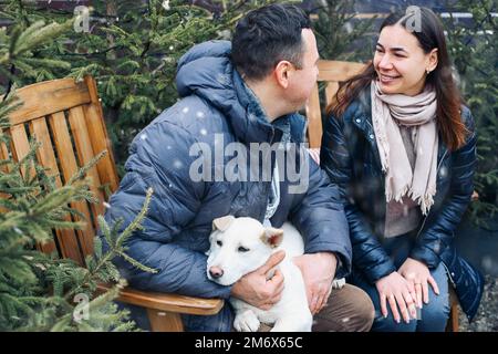 Bella giovane coppia con carino cane seduto sul cortile esterno circondato da giovani alberi di conifere Foto Stock