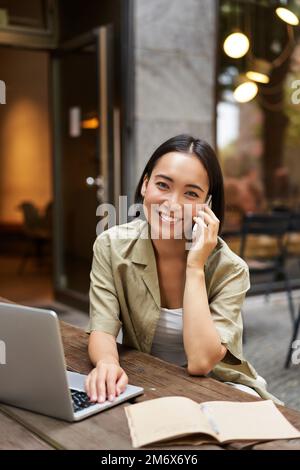 Ragazza felice che chiama sul telefono cellulare, parla sullo smartphone e lavora con il computer portatile, fa i compiti, studia all'aperto dal bar Foto Stock