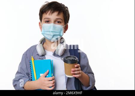 Primo piano bel ragazzo con maschera medica protettiva, tenendo fuori una tazza di carta usa e getta con bevanda calda alla macchina fotografica Foto Stock