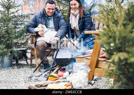 Bella coppia con cane carino seduto sul cortile esterno circondato da giovani alberi di conifere Foto Stock