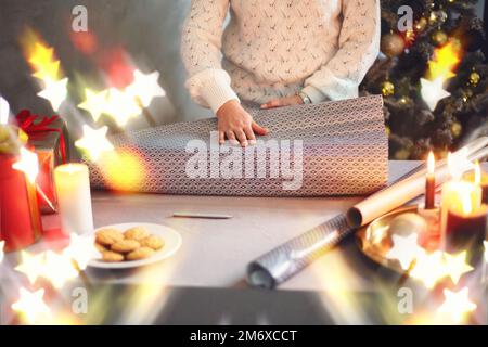 Donna di mezza età in maglione bianco che avvolge i regali di Natale per la famiglia e gli amici mentre si trova a tavola vicino bella decora Foto Stock