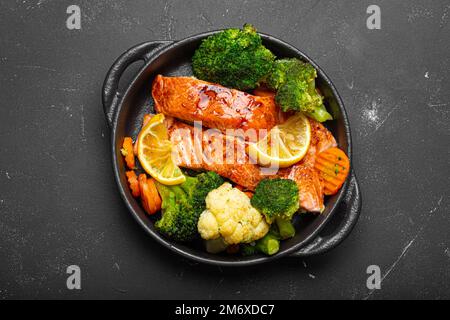 Pesce sano al forno bistecche di salmone, broccoli, cavolfiore, carota in casseruola in ghisa su fondo di pietra nera scura. Cuoco Foto Stock