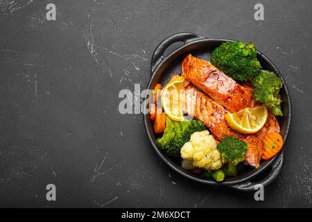 Pesce sano al forno bistecche di salmone, broccoli, cavolfiore, carota in casseruola in ghisa su fondo di pietra nera scura. Cuoco Foto Stock