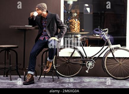 Caffè per rendere la giornata più lunga. un bel giovane che si gode una tazza di caffè in un bar della città mentre la sua bicicletta si trova accanto a lui. Foto Stock