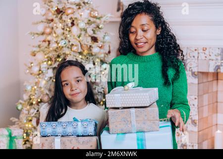 Gioiosa madre afroamericana e figlia brunetta con capelli lunghi posa contro Natale albero decorazione tenendo presenti Foto Stock