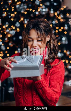 Giovane bruna donna dai capelli ricci in maglione rosso festivo si sorprende con il presente e sorride in piedi contro l'albero di Natale Foto Stock