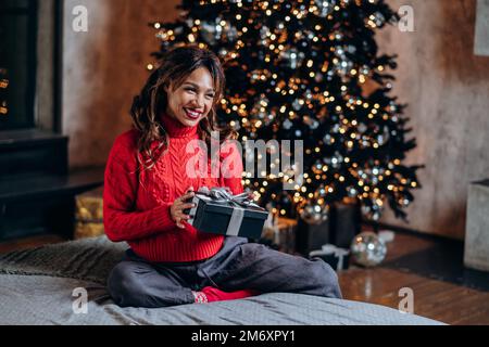 La donna dai capelli ricci bruna indossando il maglione lavorato a maglia rosso sorride brillantemente sorpreso con il presente e si siede contro l'albero di Natale con gli ornamenti Foto Stock