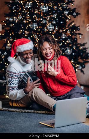 Coppia in maglioni di festa parla tramite videocall utilizzando il portatile e tiene presente seduto contro l'albero di Natale con ornamenti e ghirland Foto Stock