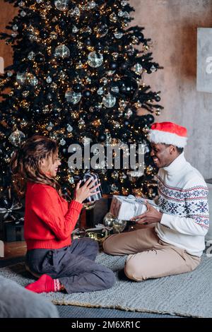 Giovane coppia felice presenta i regali l'un l'altro sorridendo ampiamente e sedendosi sul tappeto lavorato a maglia vicino all'albero di Natale decorato con ornamenti e ghirland Foto Stock