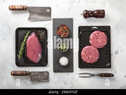 Bistecca di filetto di manzo fresco crudo e hamburger di mince sigillati in un vassoio vuoto su sfondo chiaro con utensili da barbecue. Vista dall'alto. Foto Stock