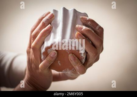 Un primo piano di mani umane sporche di argilla che tiene un piccolo vaso di ceramica bianco Foto Stock