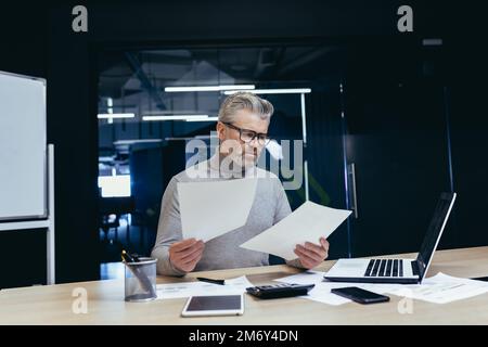 Frustrato e arrabbiato uomo d'affari con i documenti, maturo grigio uomo d'affari che controlla i rapporti finanziari, capo non soddisfatto con i risultati di realizzazione e reddito che lavora all'interno dell'ufficio con il laptop. Foto Stock