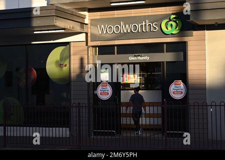 Logo del supermercato Woolworths illuminato dal sole, sopra l'ingresso del negozio, mentre un uomo cammina attraverso le porte scorrevoli del negozio Foto Stock