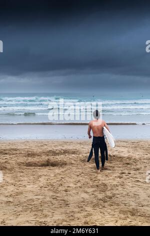 Tempo del Regno Unito; oscure nuvole di pioggia che si avvicinano come un surfista cammina verso il mare portando la sua tavola da surf a Fistral Beach a Newquay in Cornovaglia a Englan Foto Stock