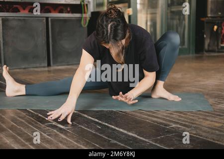 Donna yoga e nuova insegnante giovane adulto che fa uno skandasana posa in movimento per cambiare posa indossando leggings verdi durante la sua pratica di yoga su un tappetino in legno Foto Stock