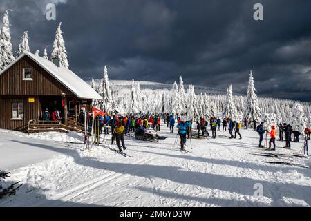 Paesaggio invernale con alberi, neve, capanna di legno, pista da sci di fondo e persone che sciano in una giornata di sole sui Monti Jizera. Foto Stock