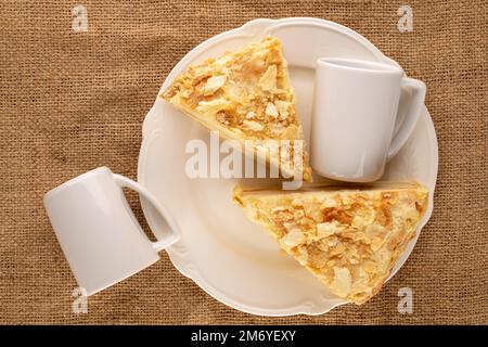 Due pezzi di torta alla crema con un piatto bianco e due tazze su un panno di iuta, macro, vista dall'alto. Foto Stock