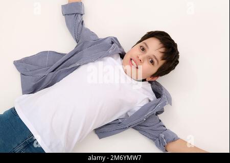 Vista dall'alto dell'adorabile ragazzo caucasico in jeans blu e t-shirt bianca, sdraiato su sfondo bianco. Spazio pubblicitario Foto Stock