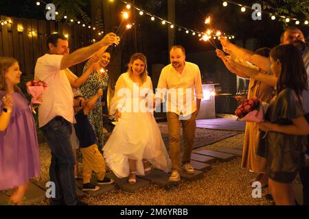 Sposate, sposate e sposate gli ospiti con luci bengala accese all'aperto durante la festa di nozze dei cocktail. Concetto di felicità, celebrazione e amore Foto Stock