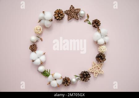 Natale inverno cornice rotonda di rami thuja, fiori di cotone, coni di abete, stelle di paglia e palle su sfondo rosa. Disposizione piatta, vista dall'alto, spazio di copia Foto Stock