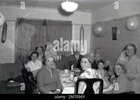 1950s, storico, partito di famiglia, sorrisi grandi come famiglia, la maggior parte che indossa i cappelli della festa, siede in una stanza anteriore intorno ad un tavolo che ha un pasto insieme, palloncini sui wallls, Inghilterra, Regno Unito. Foto Stock