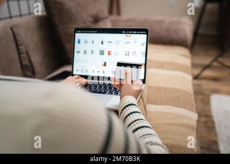 Una donna che acquista online tenere carta di credito e computer portatile a casa vista posteriore di giovane donna adulta fare l'acquisto online mentre si siede nel sof Foto Stock