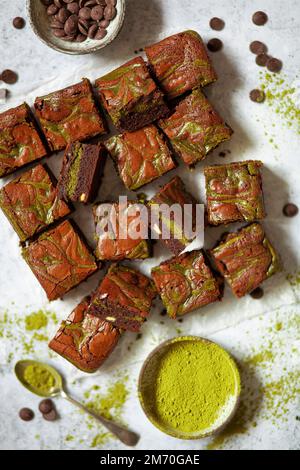 Quadrati di brownie al cioccolato con un vortice di cheesecake matcha e ciotole di polvere di matcha e patatine di cioccolato accanto. Foto Stock