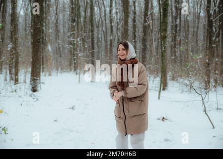 una ragazza in una foresta innevata si rallegra della neve; indossa una giacca con cappuccio di una felpa con cappuccio e una sciarpa e scarpe da ginnastica Foto Stock