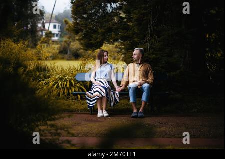 Una foto romantica di una giovane coppia seduta su una panchina in un bellissimo parco autunnale che tiene le mani, guardandosi l'un l'altro con amore e affetto Foto Stock