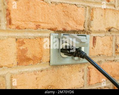 Cavo di alimentazione inserito nella presa elettrica a muro per esterni. Doppia presa di corrente coperta sulla parete in mattoni. Spina del cavo di prolunga nera. Foto Stock