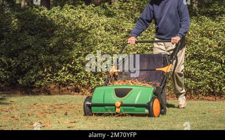 Uomo che utilizza la spazzatrice manuale per rimuovere le foglie cadute dal prato erboso del cortile residenziale. Foto Stock