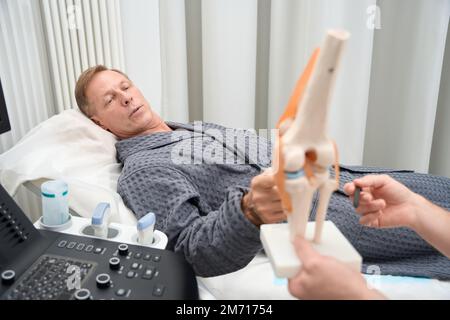 Uomo tranquillo in abito d'ospedale che indaga circa anatomia umana del ginocchio Foto Stock