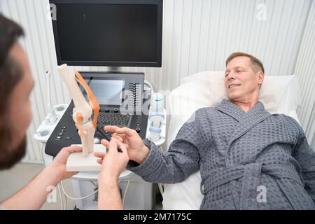 Uomo adulto in abito d'ospedale che indaga circa anatomia umana del ginocchio Foto Stock