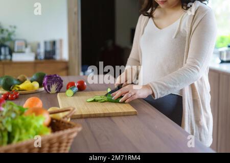 Donna asiatica incinta cuttando cetriolo per insalata verde fresca, femmina prepara gustosa cena biologica a casa, nutrizione sana per futura madre Foto Stock