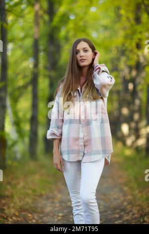 Donna bionda in abito casual a piedi lungo autunno tiglio albero vicolo. Percorso sotto gli alberi gialli con foglie autunnali in caduta. Giorno autunnale soleggiato. T Foto Stock