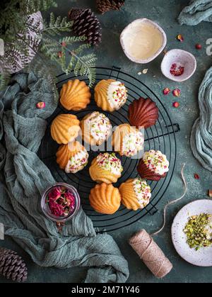 Madeleines festive su una griglia di raffreddamento decorata con cioccolato bianco, pistacchi e petali rossi su uno sfondo verde e panno Foto Stock