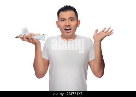 L'uomo ispanico dalla pelle scura con un volto confuso mostra il suo trattamento ortodontico, mentre tiene un dentifricio, gli spazzolini e il filo interdentale in una mano, una Foto Stock