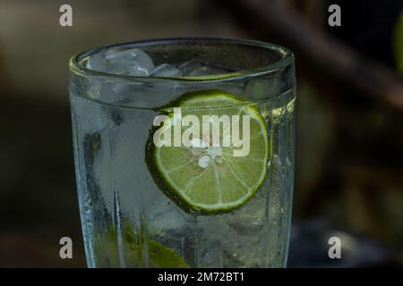 Un bicchiere d'acqua fredda al limone con cubetti di ghiaccio in un bicchiere di vetro trasparente, un caffè sfocato sfondo Foto Stock