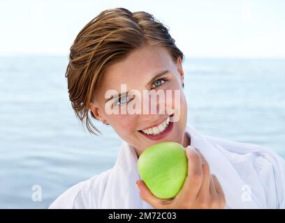 Un boccone in spiaggia. Una giovane donna attraente che prende un morso da una mela verde. Foto Stock