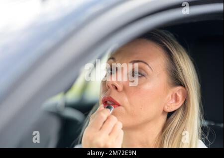 Attraente donna di mezza età bionda seduta in un'automobile che guarda nello specchio dopo l'applicazione del rossetto Foto Stock