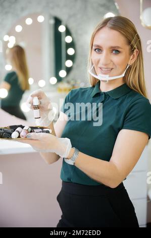 Bella giovane donna con maschera protettiva e guanti sterili che tengono una bottiglia di siero. Donna sorridente estetista che mostra prodotti cosmetici nel salone di bellezza. Foto Stock