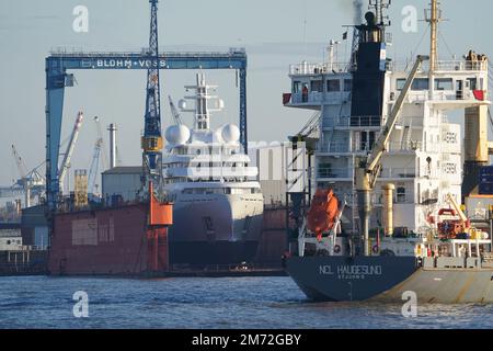 PRODUZIONE - 03 gennaio 2023, Amburgo: Una nave container naviga verso un terminal container nel porto. Sullo sfondo, uno yacht di lusso si trova in una darsena presso il cantiere navale di Blohm Voss. Il porto di Amburgo sull'Elba è un porto universale per tutti i tipi di merci e di gran lunga il più importante centro di movimentazione merci in Germania. Ogni anno, circa 130 milioni di tonnellate di merci di mare passano oltre i bordi del molo attraverso un buon 50 impianti di movimentazione, e circa 290 ormeggi offrono spazio per navi di tutte le dimensioni - da container e navi portarinfuse particolarmente grandi a petroliere e petroliere chimiche, navi di alimentazione e wat interno Foto Stock