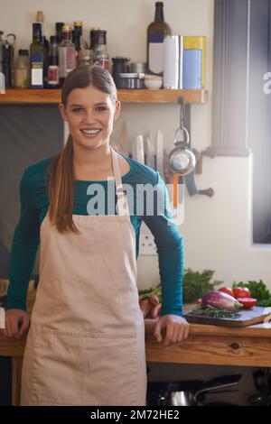 Si sente a casa in cucina. Ritratto di una giovane donna attraente che indossa un grembiule in piedi in cucina. Foto Stock